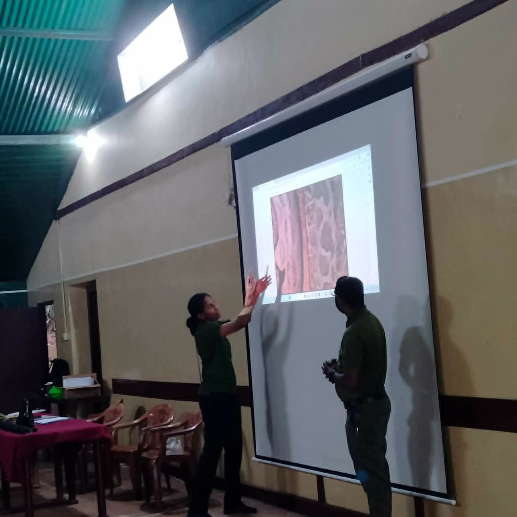 A presenter pointing at a projected close-up image during a community awareness session, with an attendee looking on